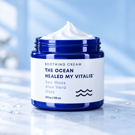A jar of soothing cream with white whipped cream on top, set on a light marble surface with a soft blue background. The jar is dark blue with a white label that reads 'SOOTHING CREAM THE OCEAN HEALED MY VITALIS Sea Moss Aloe Vera Oats'.